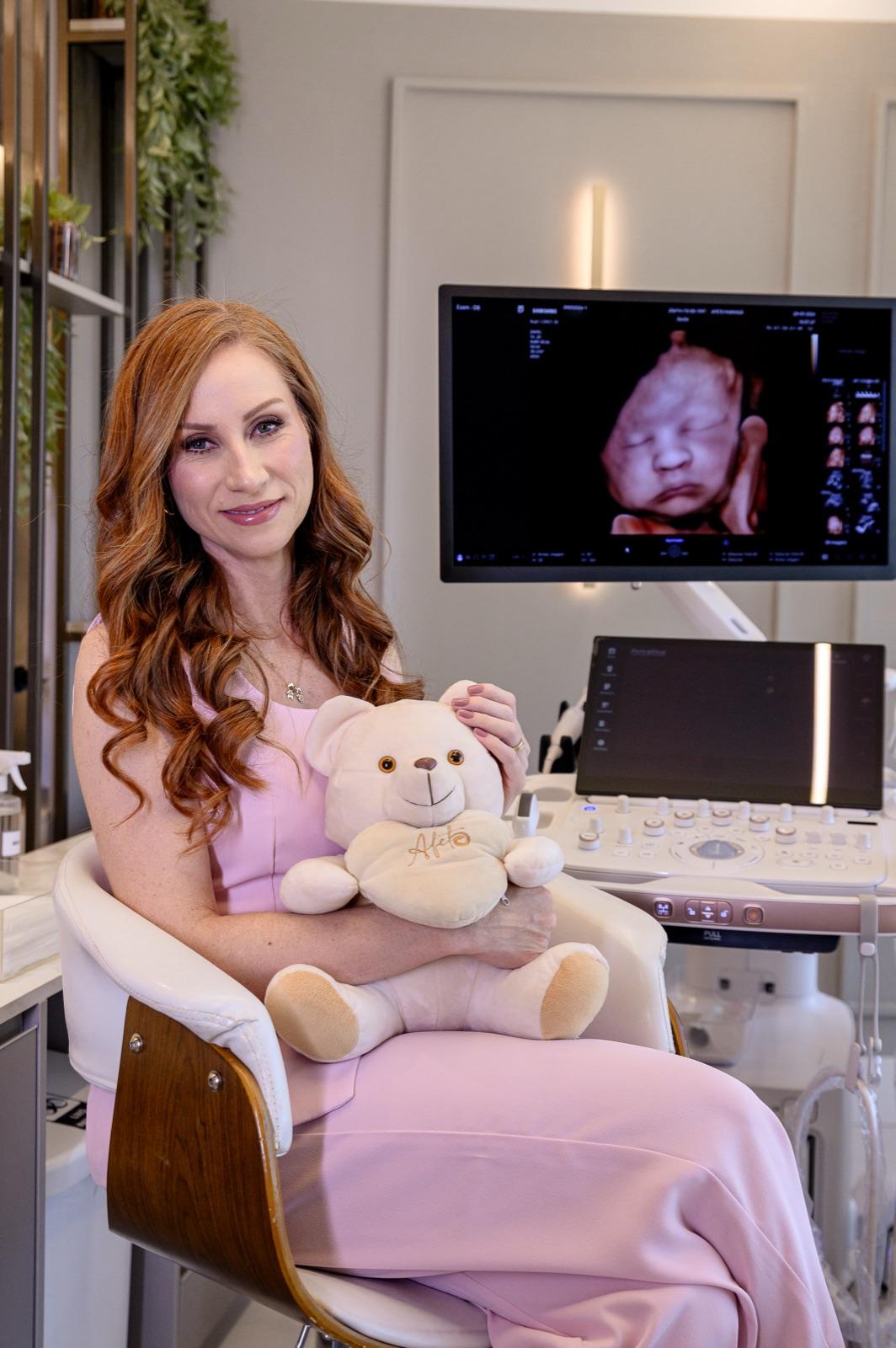 Dra. Bruna com o urso Afeto no colo, ao lado do aparelho de ultrassom com a imagem do bebê em 8D na tela